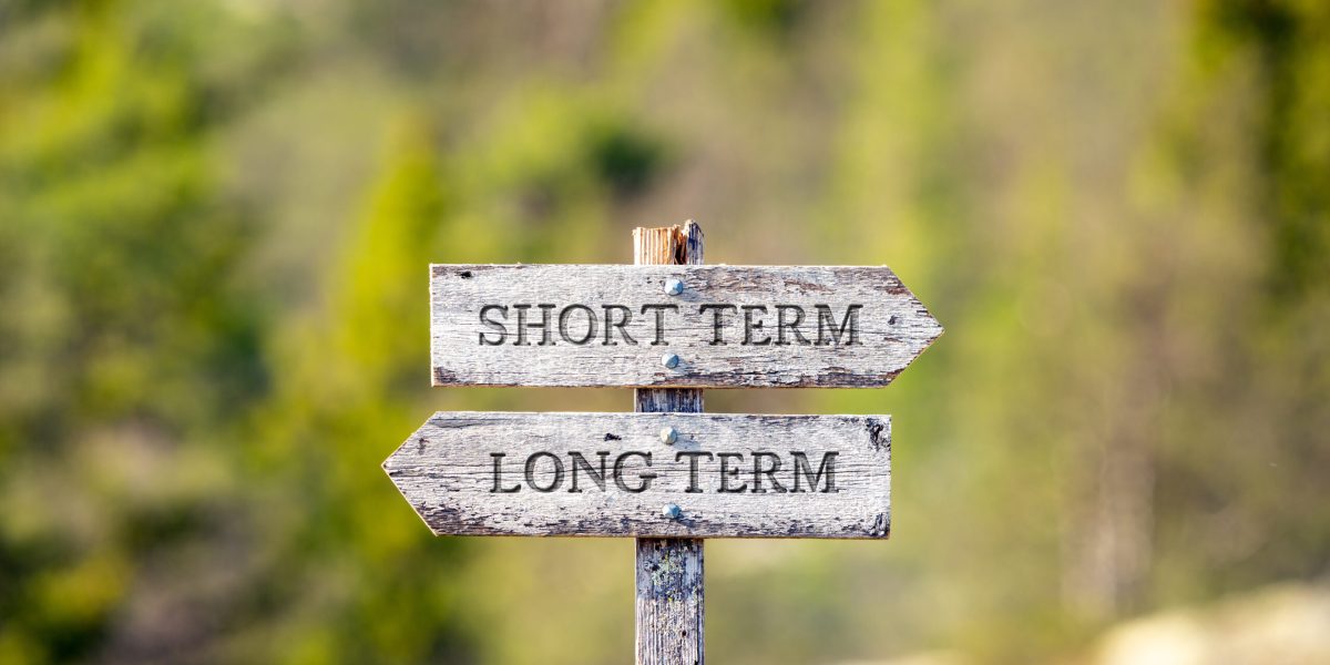 short term long term text carved on wooden signpost outdoors in nature. Green soft forest bokeh in the background.